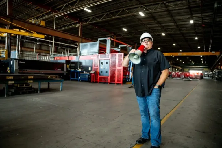 Man wearing a white hard hat and safety glasses holds a red megaphone while standing in a large industrial warehouse.