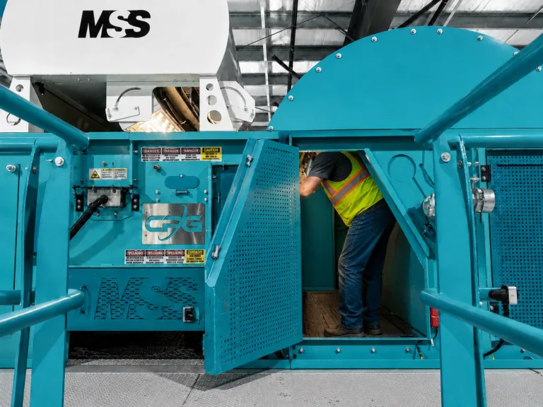 Worker in neon safety vest and jeans leaning into an open turquoise industrial machine with perforated door and MSS logo.