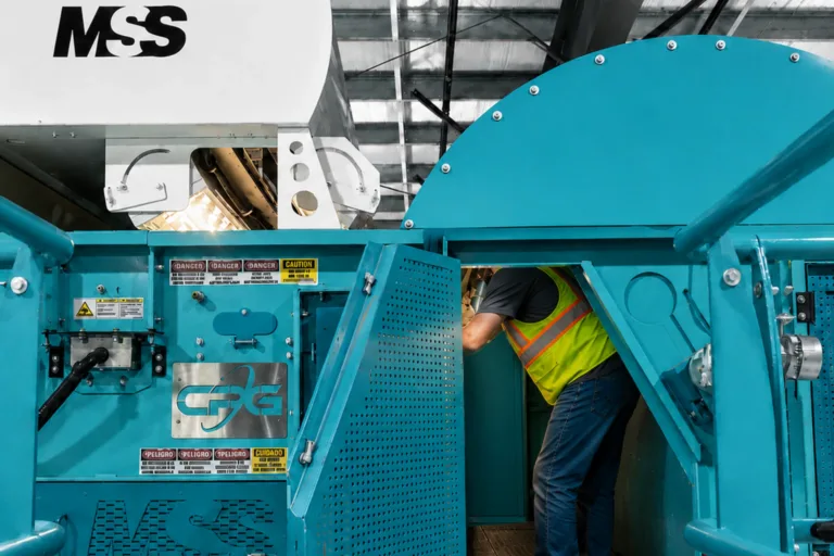 Worker in yellow safety vest leaning into open teal industrial machine with MSS logo and warning labels.