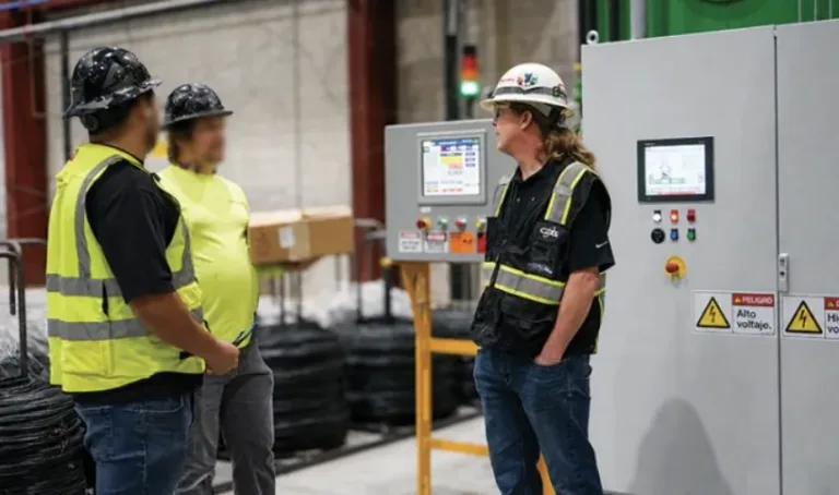 Three workers in hard hats and high-visibility vests standing beside industrial control panels with electrical warning signs