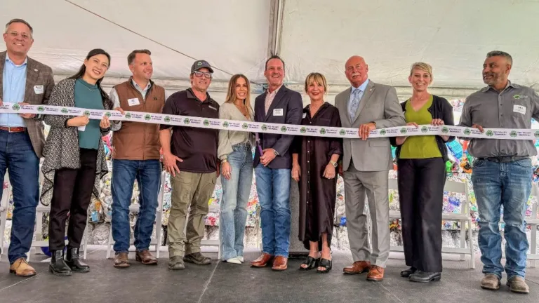 Ten people stand in a row under a tent holding a long ribbon for a ribbon-cutting ceremony.