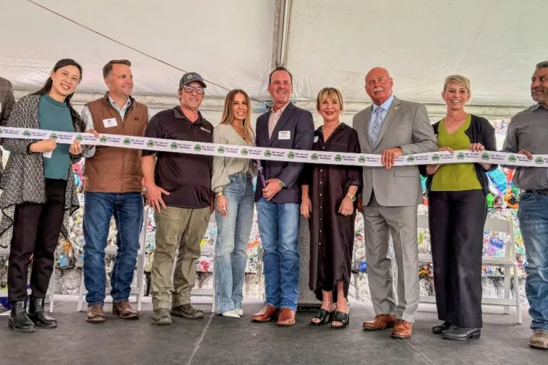 Ten people stand in a row under a tent holding a long ribbon for a ribbon-cutting ceremony.